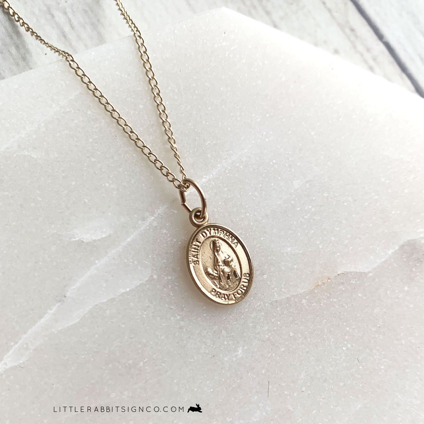 Saint Dymphna is the patron saint of those suffering from mental illness, mental health disorders, anxiety and depression. She is a catholic saint. This St. Dymphna gold necklace is delicate and dainty. It's made of 14kt gold filled material and suspended from a delicate gold chain. Pictured here on a white stone background. This necklace would be the perfect catholic confirmation gift for her.