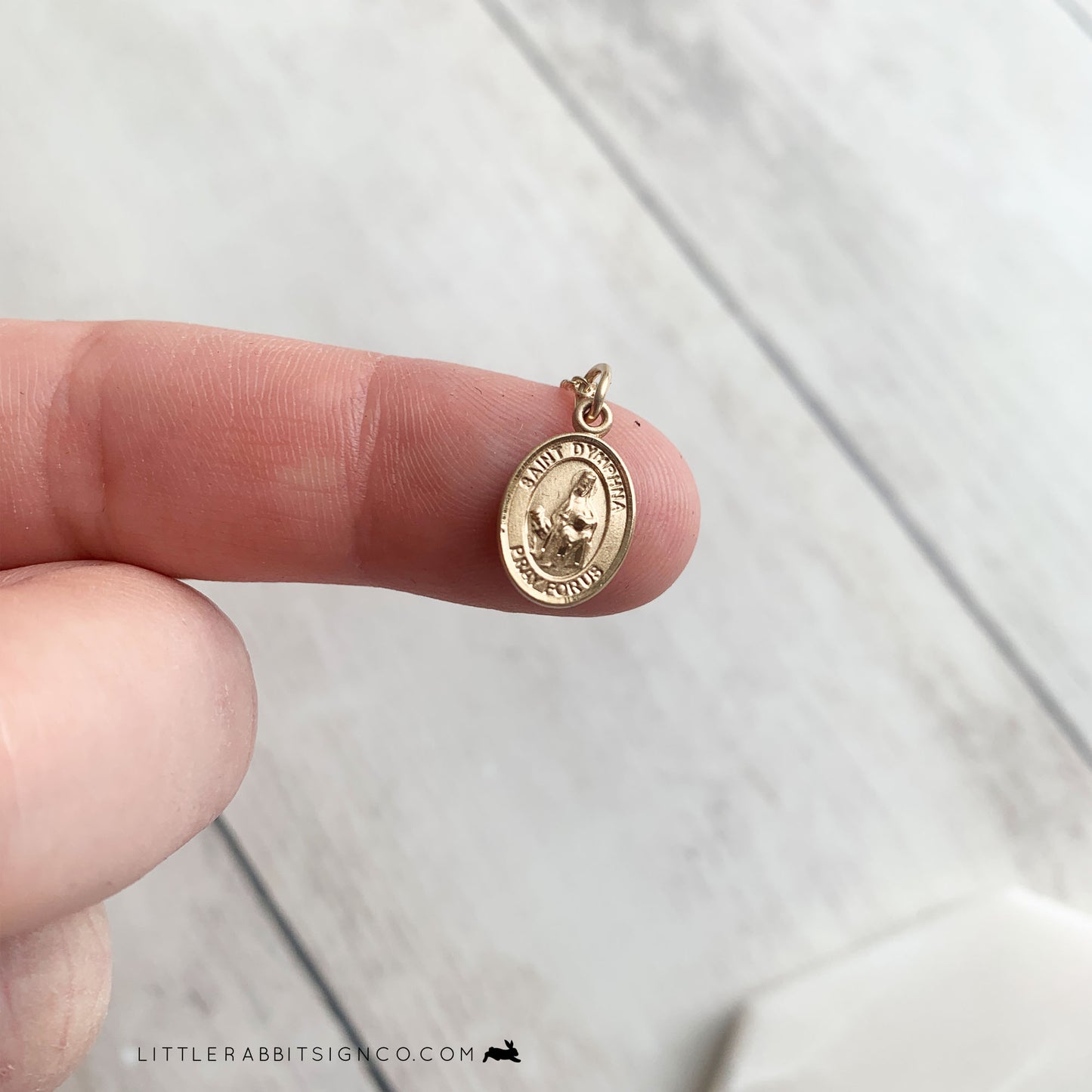 Tiny gold Saint Dymphna medal is shown on the fingertip of a person. This image depicts the small size of the medal. St Dymphna is the patron saint for those suffering from anxiety, depression, and mental illness, and is a powerful saint in the Catholic Church.