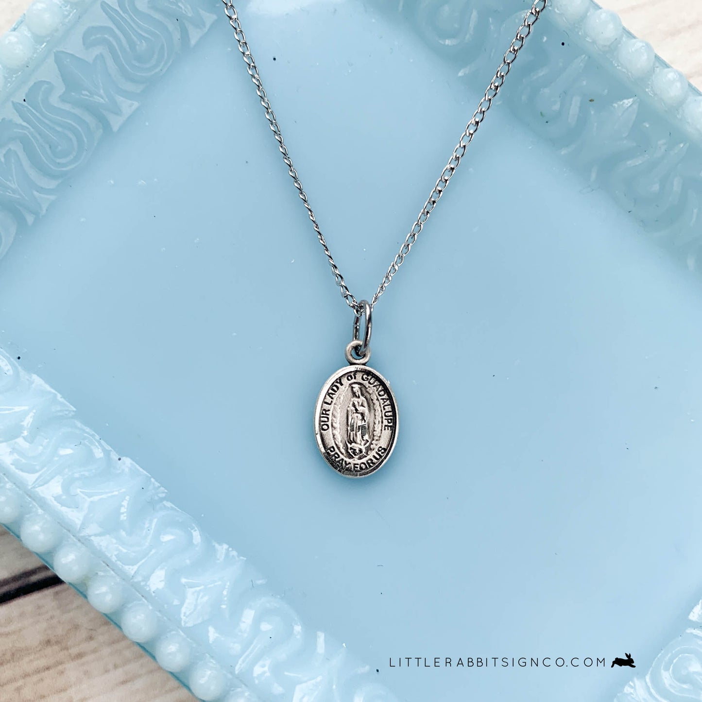 Pictured is a dainty Our Lady of Guadalupe sterling silver medal suspended on a sterling silver light curb chain with a blue background. Our Lady of Guadalupe Feast Day is December 12. OLG Pray for Us.