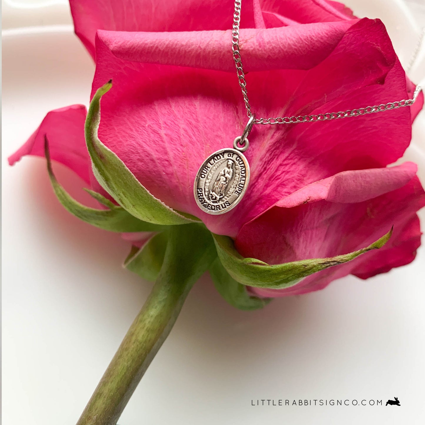 Beautiful Our Lady of Guadalupe necklace in sterling silver is shown laid upon a pink rose in this image.