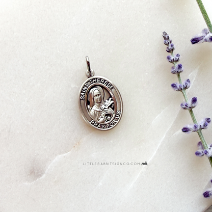 Silver Saint Therese pendant on a white fabric background with lavender flowers.